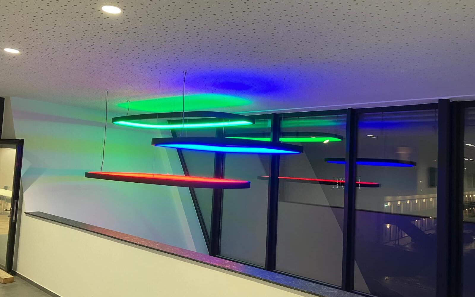 SOFT SQUARE mit RGB-Licht - Rechteckige Ringpendelleuchten im Treppenhaus Blick von einem Büro Balkonflur der KSK-Bau Freiburg auf mehrere rechteckige großformatige Ringleuchten SOFT SQUARE mit schwarzem Profil, die in den verschiedenen RGB-Farben leuchten und in verschiedenen Höhen von der Decke abgehangen sind.