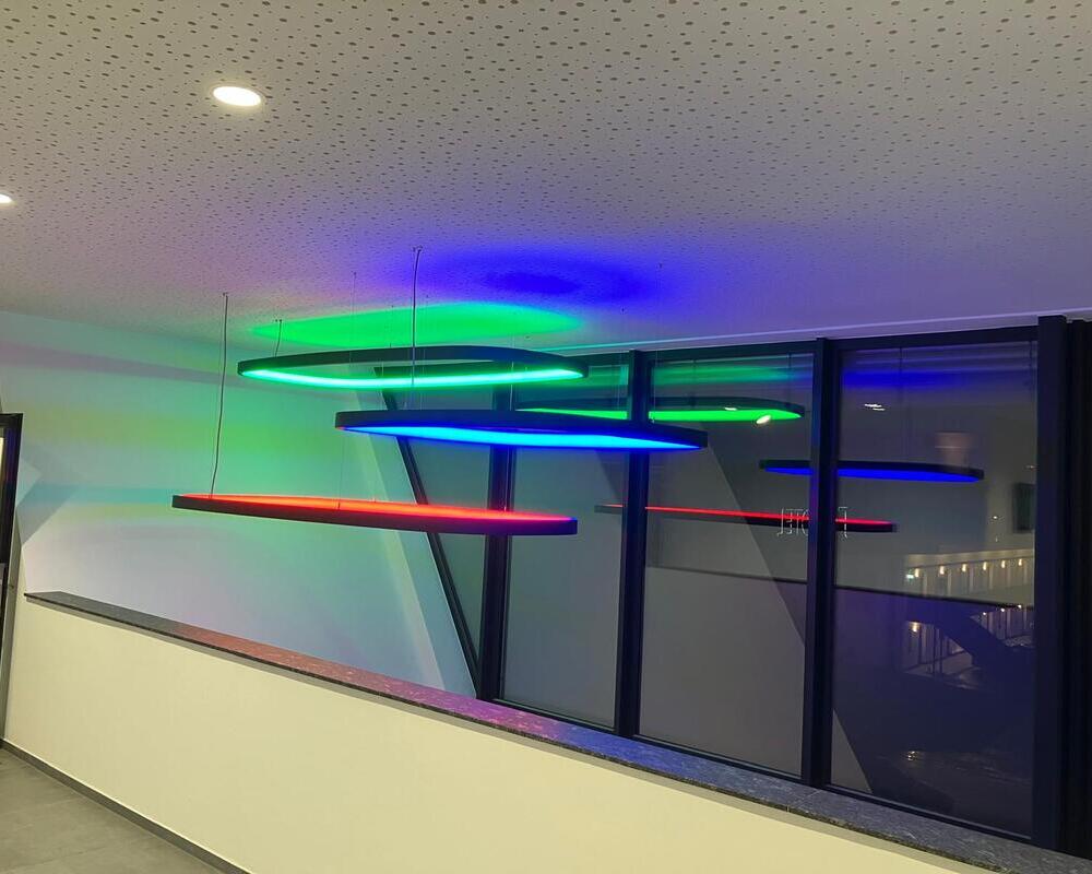 SOFT SQUARE with RGB light - Rectangular ring pendant luminaires in stairwells View from an office balcony corridor at KSK-Bau Freiburg of several rectangular, large-format SOFT SQUARE ring luminaires with a black profile, which light up in various RGB colours and are suspended from the ceiling at different heights.