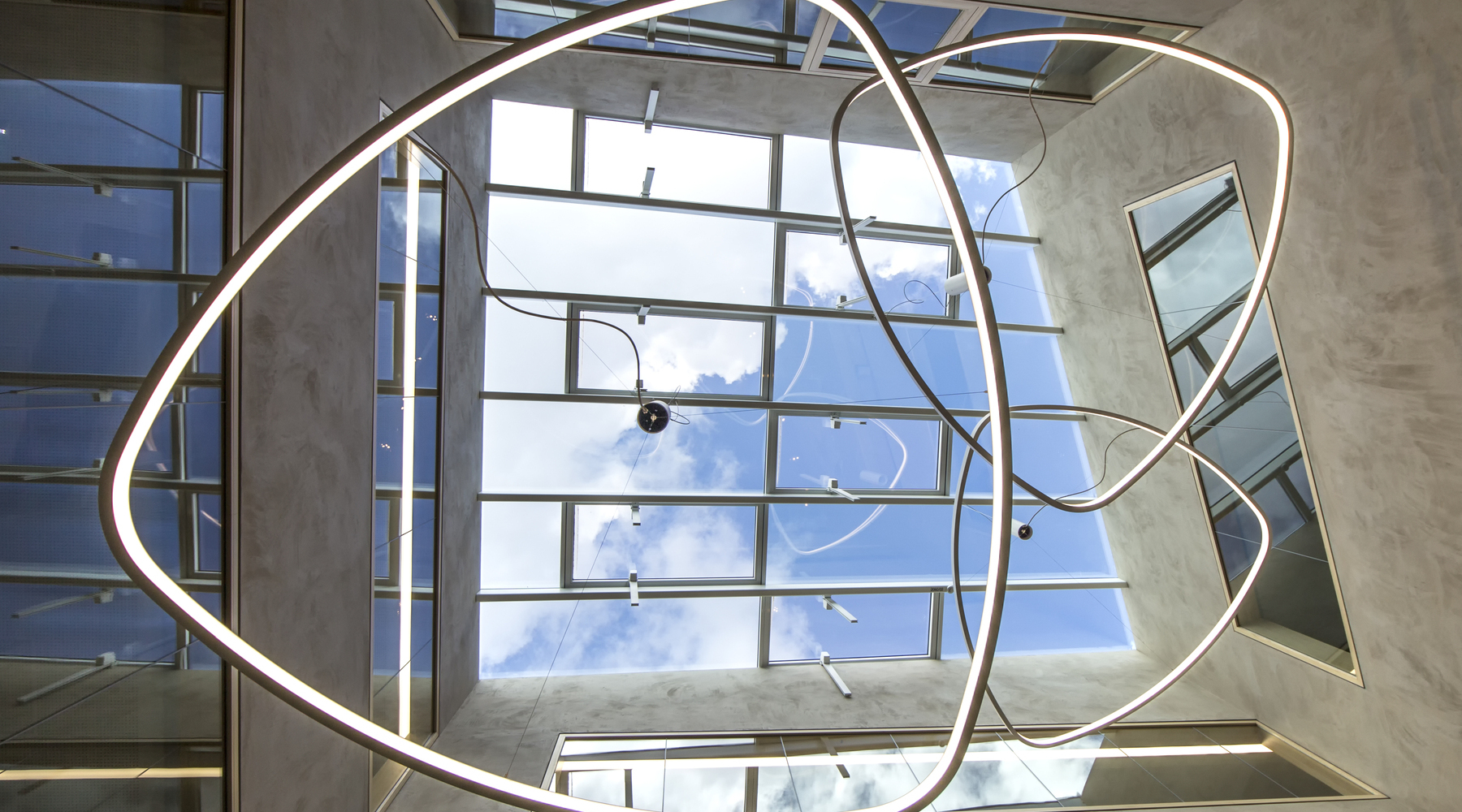 Zentraler Blick von unten in SATTLER SOFT DELTA I mit Pendelbaldachin unter Glasdach gependelt in Lobby Zentraler Blick unter der Leuchte SOFT DELTA I von SATTLER an einem Pendelbaldachin unter einem Glasdach montiert,