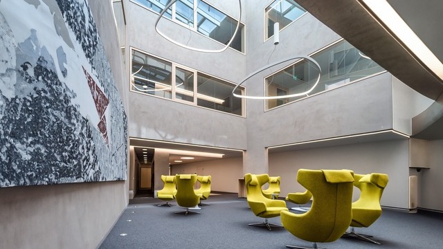 Foyer lighting in a bank. Solid-coloured, bright, comfortable upholstered wing chairs. Surrounded by light-coloured exposed concrete, an angular open space forms above. Looking further upwards, the eye glides over rounded triangular shapes: SOFT DELTA ring lights made of white powder-coated aluminium. | © SATTLER