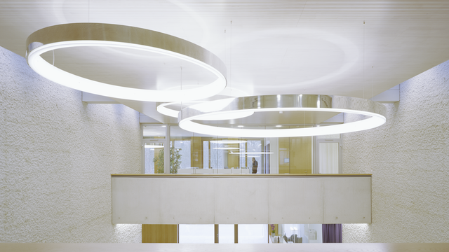 Three large-format CIRCOLO ring luminaires in polished aluminium as a focal point in the exposed concrete building combined with light-coloured wooden elements from Karl Köhler GmbH. Suspended close to the ceiling at 1st floor level of the glazed office. Free space under the luminaires can be used as an event room. | © Brigida González