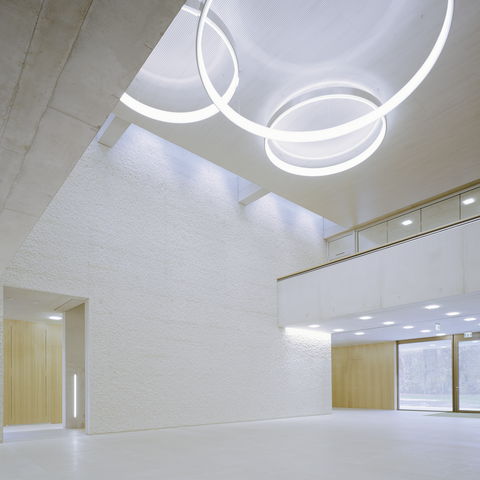 Blick von unten an die Decke eines Sichtbetongebäudes, mittig im zweigeschossigen hohen Raum hängen auf freier Fläche 3 Ringleuchten CIRCOLO in Aluminium poliert, auf beiden Seiten verengt sich der Raum um ein Stockwerk.