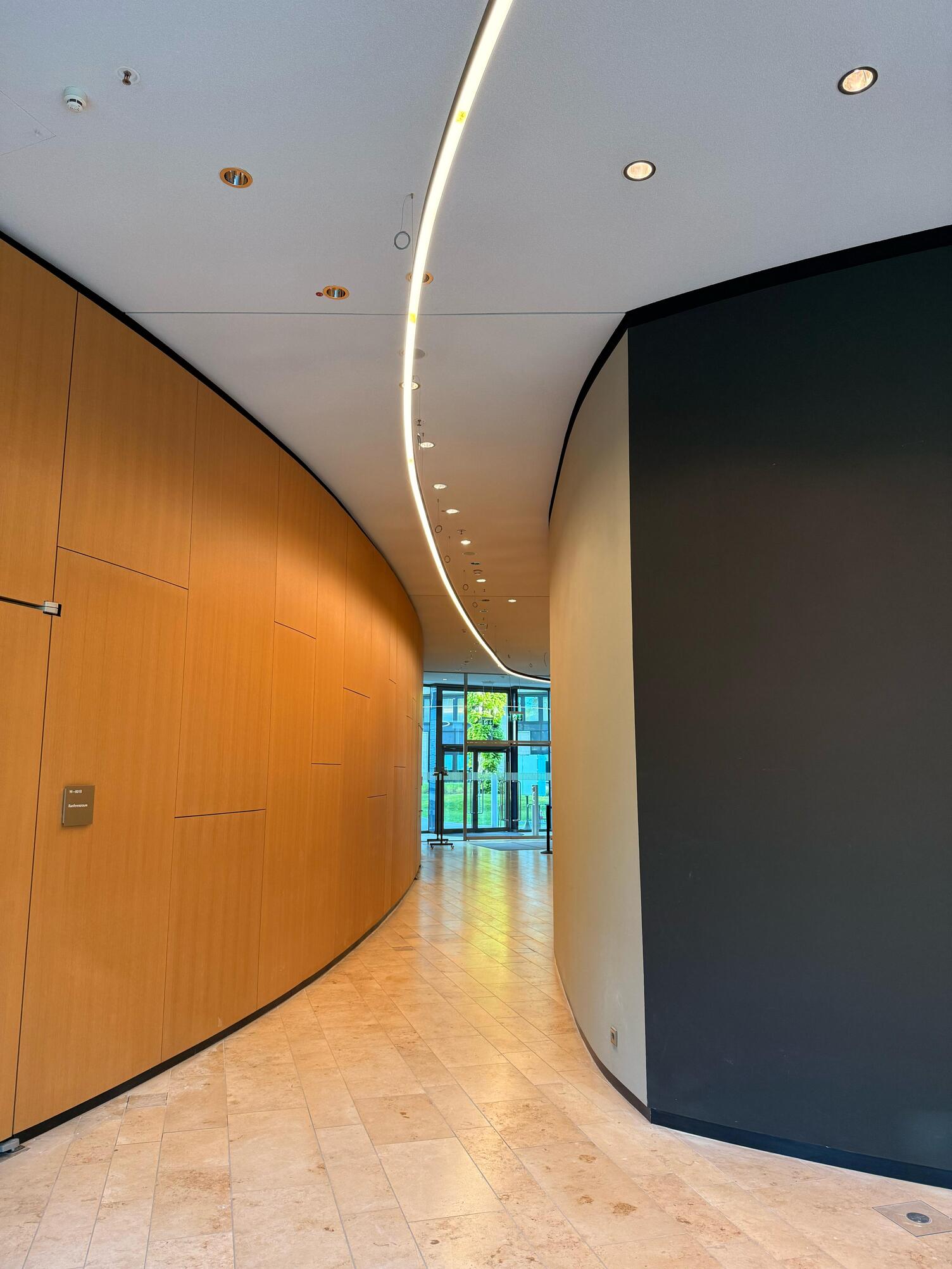 Flurbeleuchtung mit gebogener DUETTO Lichtlinie Flur in den KfW Westarkaden Frankfurt mit runden Wänden, links Holzoptik, rechts Beton, mittig an der Decke verläuft eine gebogene DUETTO Lichtlinie entlang des gebogenen Gangs.
