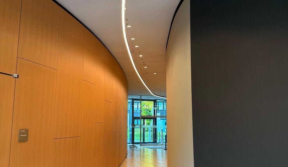 Flurbeleuchtung mit gebogener DUETTO Lichtlinie Flur in den KfW Westarkaden Frankfurt mit runden Wänden, links Holzoptik, rechts Beton, mittig an der Decke verläuft eine gebogene DUETTO Lichtlinie entlang des gebogenen Gangs.