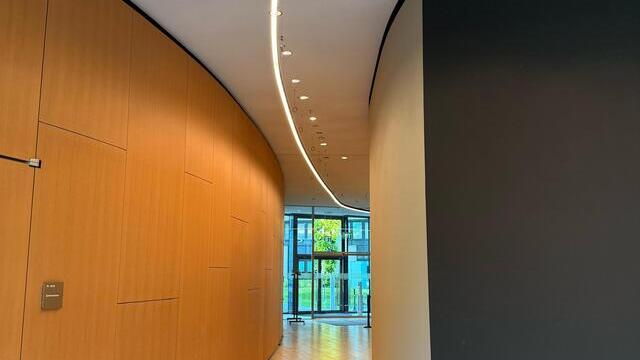 Flur in den KfW Westarkaden Frankfurt mit runden Wänden, links Holzoptik, rechts Beton, mittig an der Decke verläuft eine gebogene DUETTO Lichtlinie entlang des gebogenen Gangs. | © SATTLER