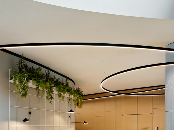 Moderner Lounge-Bereich der kfW Westarkade, links im Bild: halbrunde Treppenholzbank, grünen Kissen, Pflanzen deckennah, rechts im Bild verschwommen eine laufende Person. Oben im Bild: zwei schwarze gebogene DUETTO Lichtlinien, die von der Decke abgependelt sind und um die Säule verlaufen.  | © @Nina Struve for VRAI interior architecture