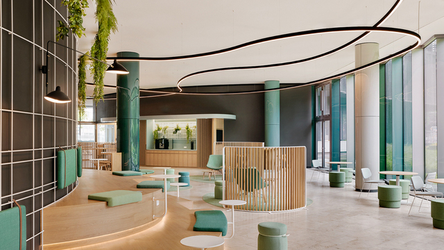 Moderner Lounge-Bereich mit grünen runden Säulen, halbrunder Treppenholzbank, grünen Kissen, Pflanzen deckennah und gebogenen schwarzen DUETTO Lichtlinien, die teils um die Säulen verlaufen. | © Nina Struve for VRAI interior architecture