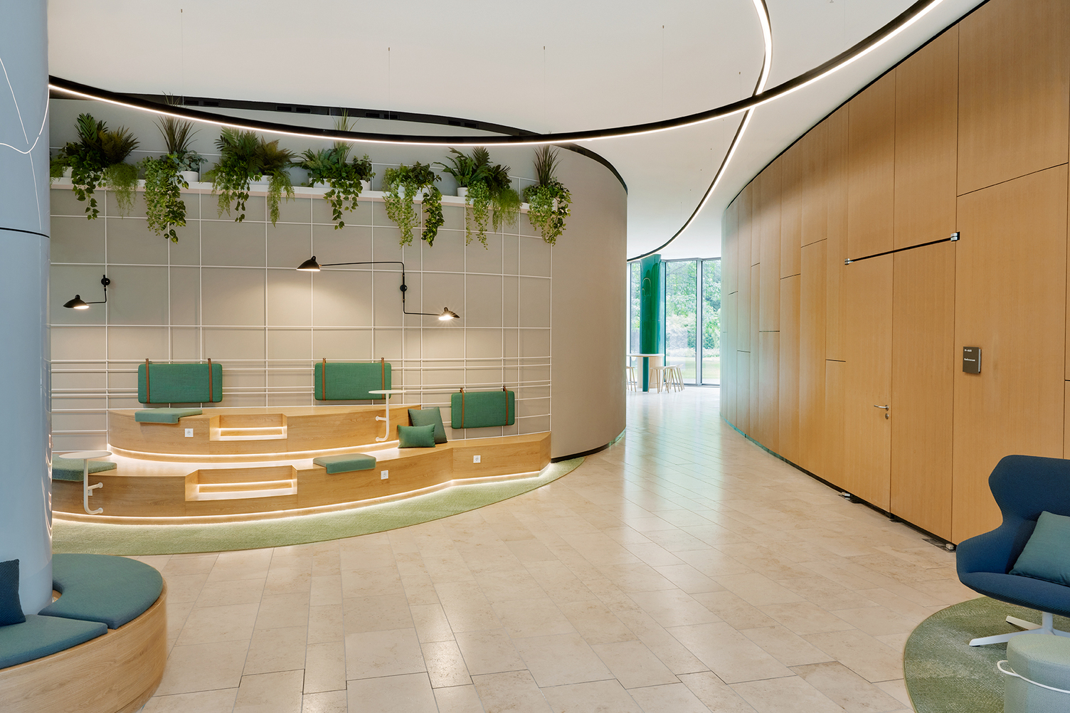 Wegweisende DUETTO Lichtlinien Moderner Lounge-Bereich der kfW Westarkade. Blick auf halbrunde zweistufige Treppenholzbank mit grünen Kissen, darüber entlang der runden Wand grüne Pflanzen. Rechts im Bild führt ein Gang zu einem weiteren Raum mit bodennahen Fenstern und Blick ins Grün. Oben im Bild: zwei gebogenen schwarze DUETTO Lichtlinien, die von der Decke abgependelt sind und auf unterschiedlichen Höhen verlaufen und sich einmal kreuzen. Eine verläuft weiter den Gang entlang.