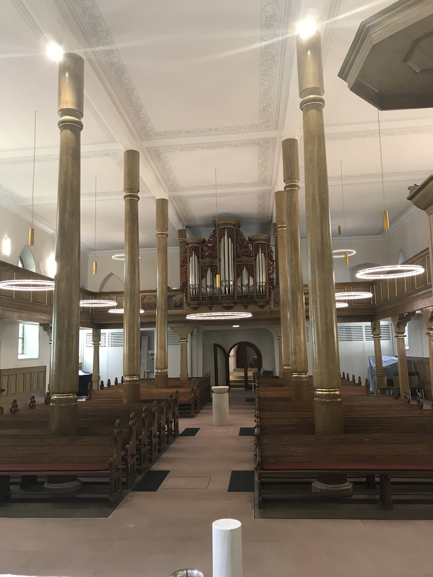 Goldene Ringleuchten für evangelische Kirche Altargang und Blick auf Kirchenorgel in der historischen Kirche in Westhofen. Hohe Raumhöhen und von Säulen durchzogener Raum, dazwischen Kirchenbänke. Von der Decke abgependelt mit einem goldenen zylindrischen Pendelbaldachin mehrere Ringleuchten in Gold, DOPPIO, direkt und indirekt leuchtend.