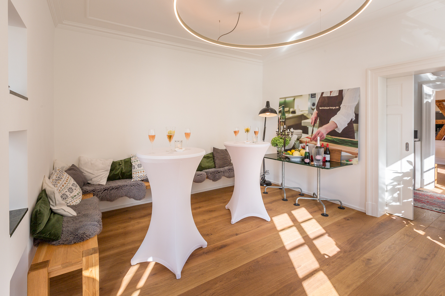Standing reception under DOPPIO ring light White room with white bar tables topped with champagne glasses, cosy wooden benches with cushions along the wall, a large round ring light suspended above the tables providing direct and indirect lighting (DOPPIO), partially visible in the image. A welcoming, friendly colour photo of a chef wearing a black apron is prominently displayed on one side wall.
