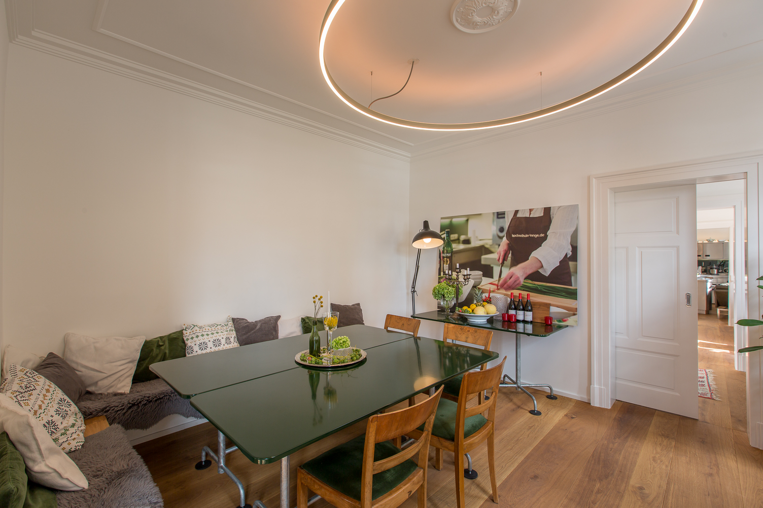 White room with green glass table, cosy wooden benches with cushions along the wall and around the table, large round ring light suspended above the tables providing direct and indirect lighting (DOPPIO), partially visible in the picture. Very prominent on one side wall is an inviting, friendly colour photo of a chef wearing a black apron.