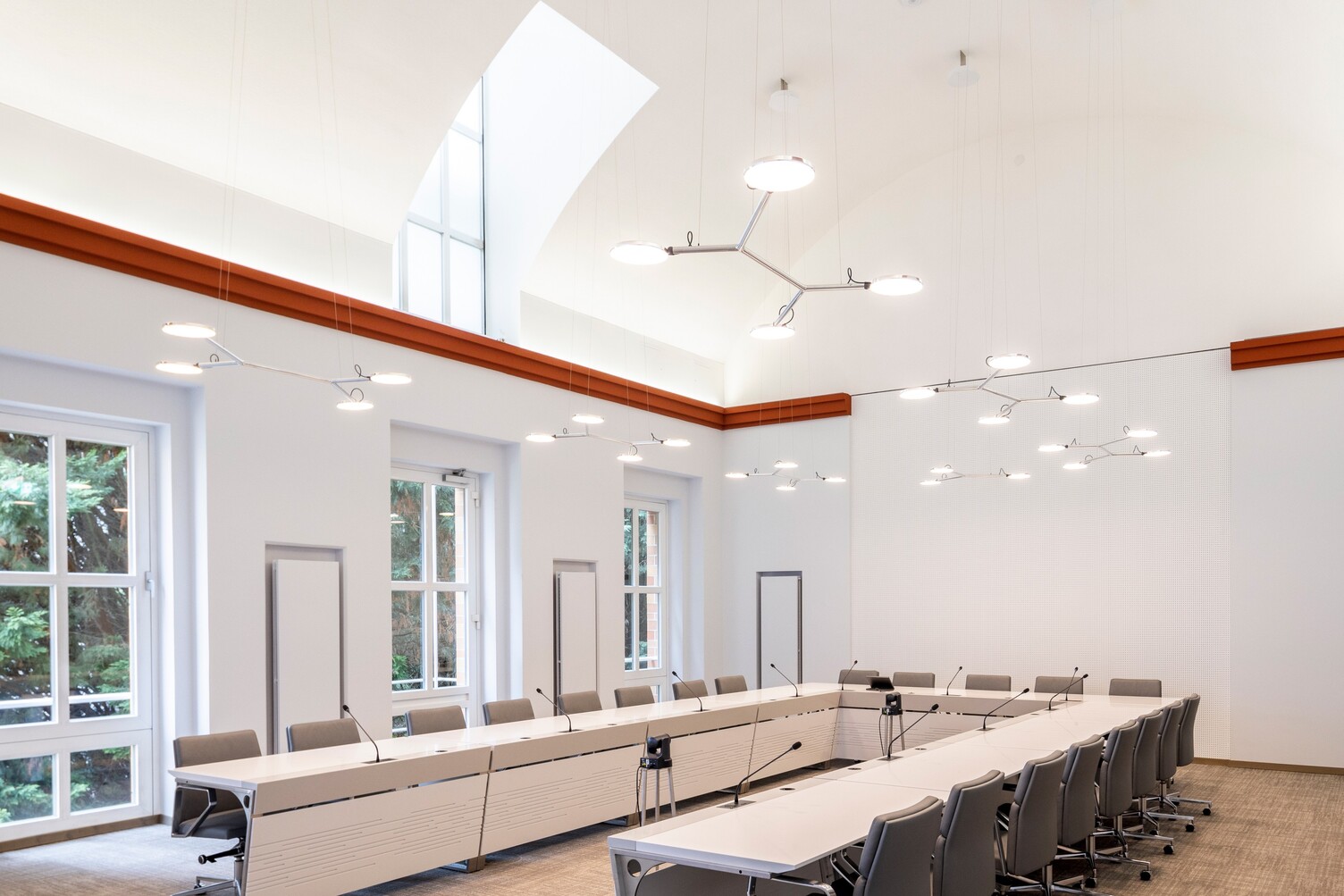 View of the front end of U-shaped seating in the Lambersart Kreis Viersen meeting room with white walls and light red, straight decorative moulding near the ceiling. Along the U-shaped table above, branched pendant luminaires with 3 to 4 light heads each (AVVENI PENDANT 3 or 4). Two single-head pendant luminaires AVVENI PENDANT 1 in front of the screen.