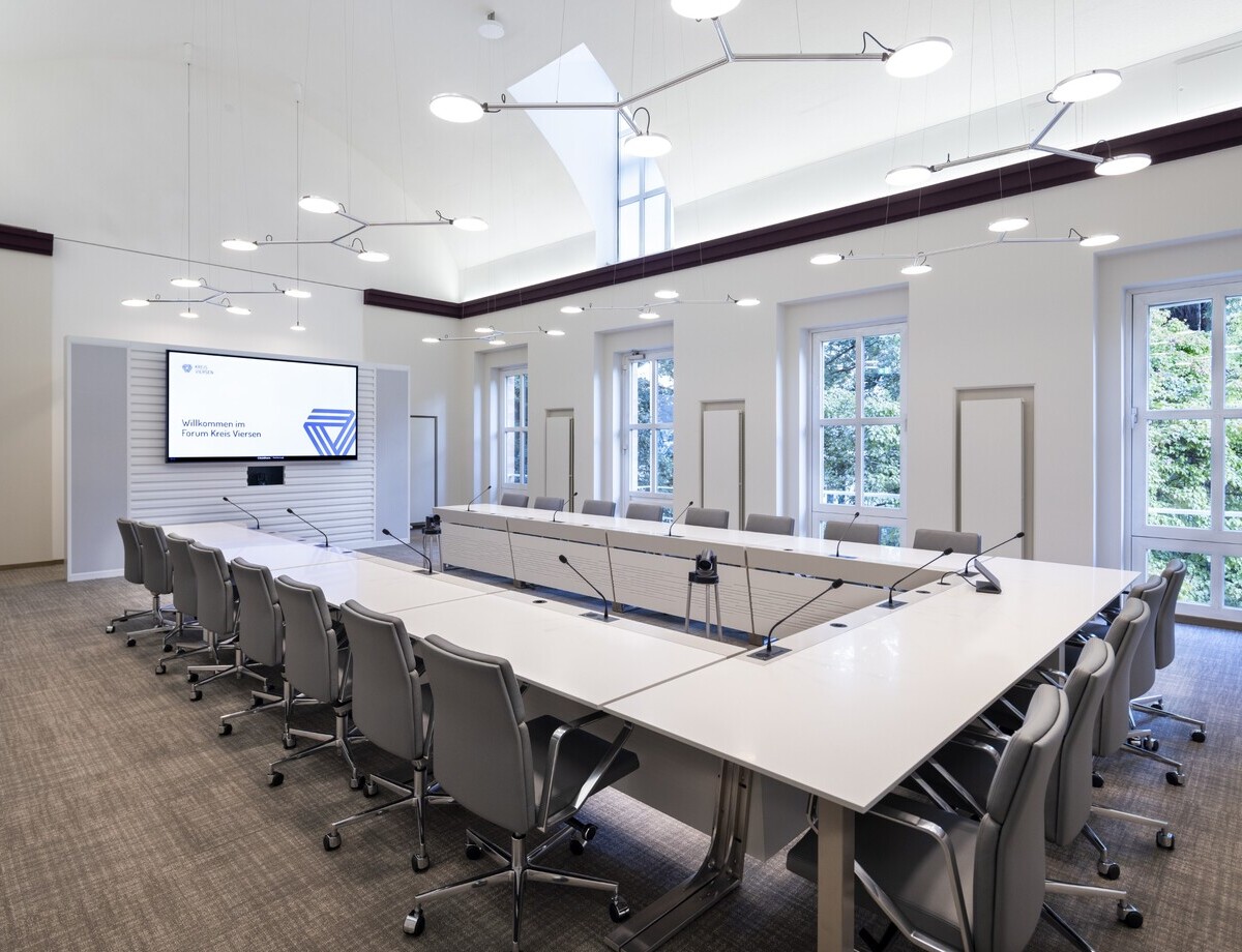 Beleuchtung AVVENI PENDANT im Sitzungsraum U-Bestuhlung Schräger Blick von einer Ecke der U-Bestuhlung im hellen Sitzungszimmer Peterborough Kreis Viersen, gestaltet von DIELEN INNENARCHITEKTEN BDIA, mit weißen Wänden und dunkelroter geradliniger deckennaher Zierleiste, auf die Fensterseite & den Bildschirm. Entlang des U-Tisches darüber verzweigte Pendelleuchten mit je 3 bis 4 Leuchtköpfen (AVVENI PENDANT 3 oder 4). Zwei Einzelkopfpendelleuchten AVVENI PENDANT 1 vor dem Bildschirm.