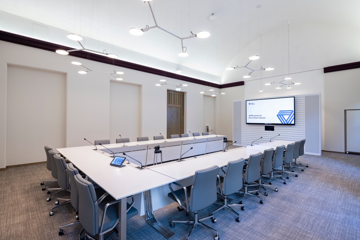 Beleuchtung AVVENI PENDANT im Sitzungsraum U-Bestuhlung Schräger Blick von einer Ecke der U-Bestuhlung im hellen Sitzungszimmer Peterborough Kreis Viersen, gestaltet von DIELEN INNENARCHITEKTEN BDIA, mit weißen Wänden und dunkelroter geradliniger deckennaher Zierleiste, auf die Wandseite und den Bildschirm. Entlang des U-Tisches darüber verzweigte Pendelleuchten mit je 3 bis 4 Leuchtköpfen (AVVENI PENDANT 3 oder 4). Zwei Einzelkopfpendelleuchten AVVENI PENDANT 1 vor dem Bildschirm.