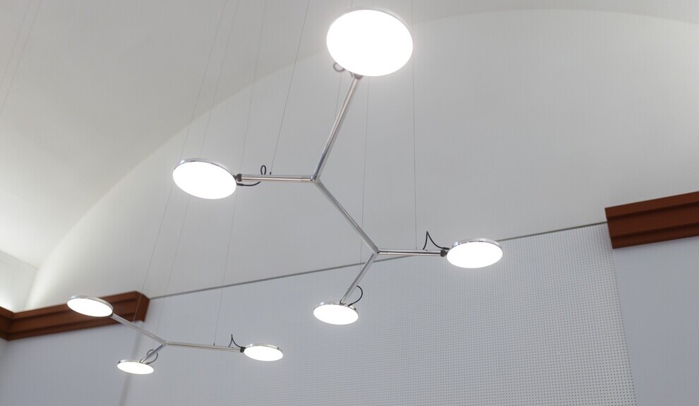 Verzweigte Pendelleuchte AVVENI PENDANT 3 & 4 Blick von unten auf 2 verzweigte Pendelleuchten vor einer weißen Wand mit abgesetzter roter Zierleiste. Eine Leuchte hat 3 runde flache Leuchtenköpfe (AVVENI PENDANT 3), eine weitere versetzt hängende hat 4 Leuchtenköpfe (AVVENI PENDANT 4).