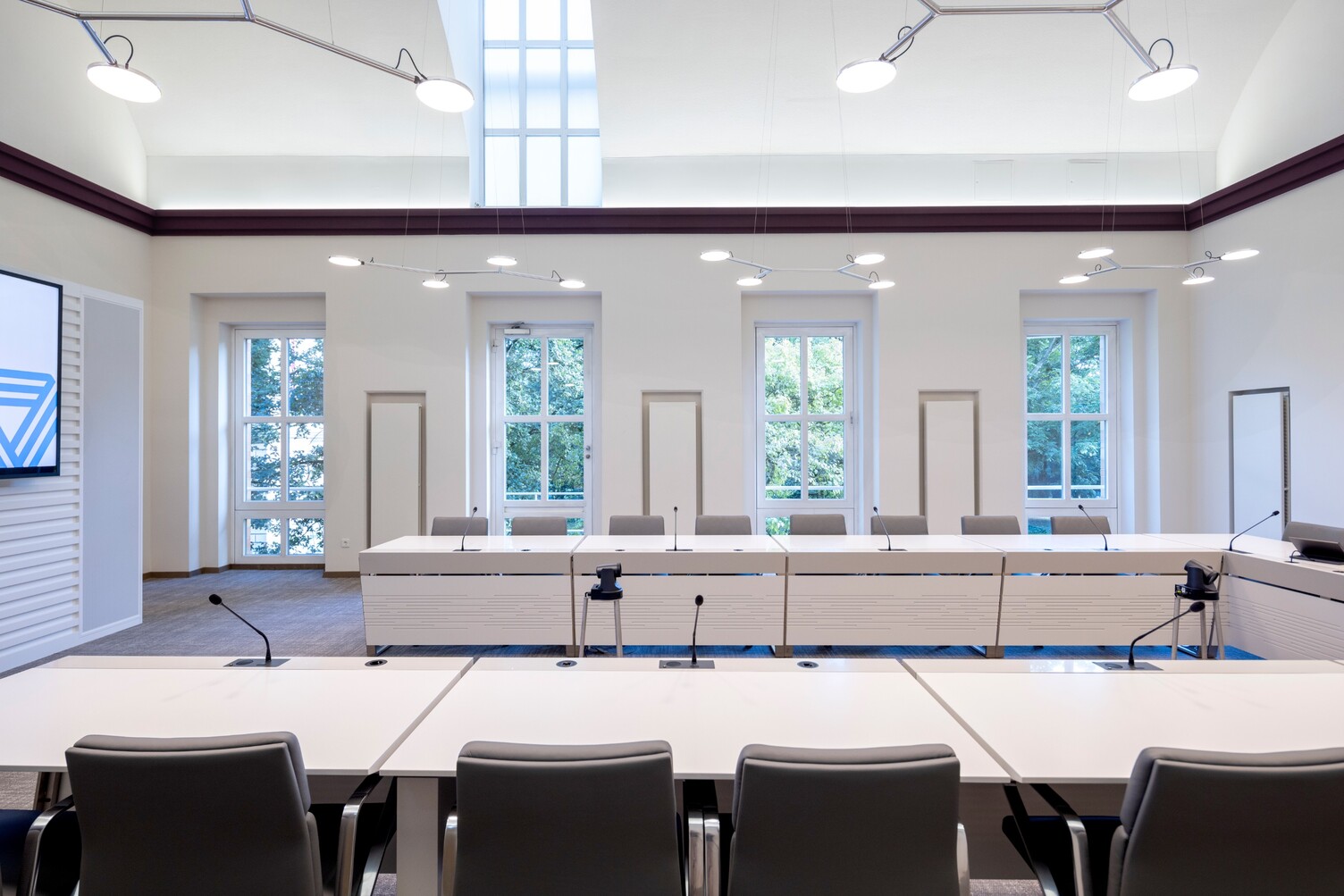 View from one long side of the U-shaped seating in the bright meeting room in Peterborough, Viersen district, designed by DIELEN INNENARCHITEKTEN BDIA, with white walls and dark red straight decorative moulding near the ceiling, towards the opposite window side. Along the U-shaped table above, branched pendant luminaires with 3 to 4 light heads each (AVVENI PENDANT 3 or 4).