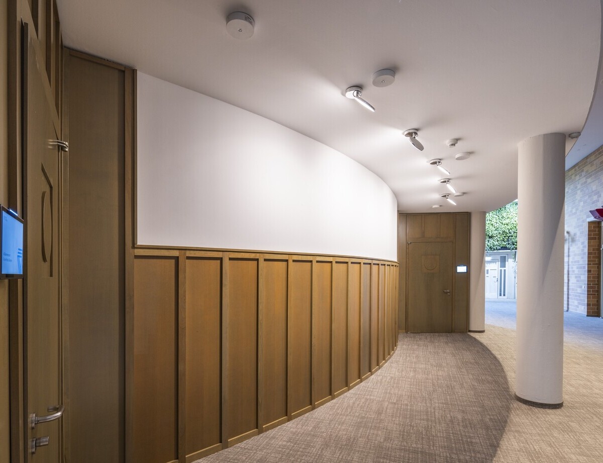 Fluraufhellung mit Anbaustrahler AVVENI SINGLE Kurvig verlaufender Flur im Forum des Kreis Viersen. Auf der einen Seite Klinkerwände, auf der anderen Wand, unten Holzverkleidet oben weiß gestrichen. An der abgehängten Decke, welche von einer weißen runden Säule gestützt ist, in Reihe mehrere AVVENI SINGLE Anbauleuchten mit runden beweglichen Kopf zur Aufhellung der Wände.