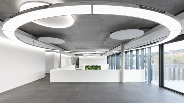 A large 7m Anello luminaire from SATTLER in a modern foyer with an exposed concrete ceiling around a concrete column, but centred on a reception desk. The lighting is complemented by acoustically effective Acoustic Light luminaires, also from SATTLER | © SATTLER | Nikolay Kazakov niko design