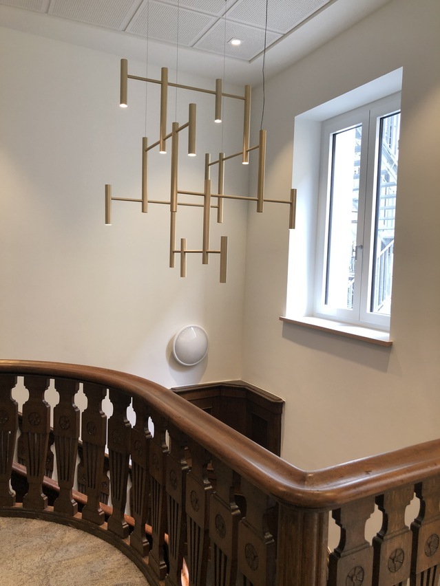 Blick über ein historisches Holzgeländer auf einen modernen rohrförmigen Kronleuchter in edlem dezenten Champagner Gold, der über der Treppe abgependelt ist.