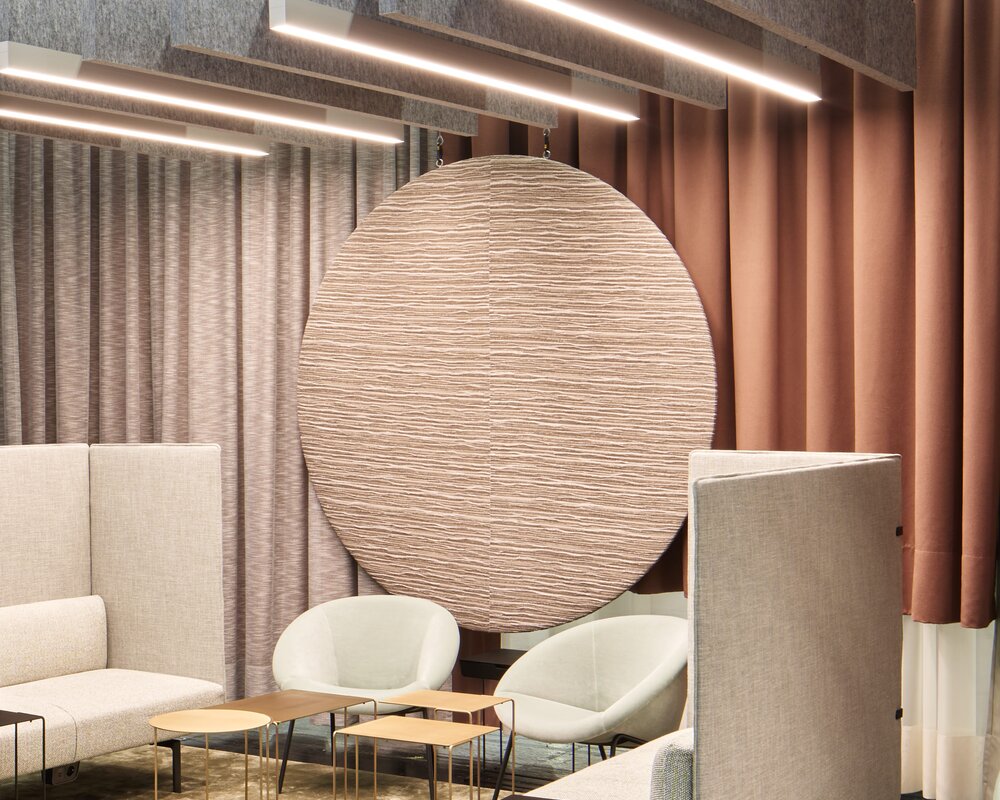 Akustisch wirksame Raum-in-Raum-Lösung mit BAFFELLEUCHTEN | © Philip Kottlorz Messestand ORGATEC 2025 von Walter Knoll mit hellen Polstermöbeln und kleinen Sofa-Tischen, darüber sind mehrere lineare Baffelleuchten (BAFFELLEUCHTE) und reine lineare akustisch wirksame Baffeln (ACOUSTIC BAFFLE) in grau abgependelt. | © Philip Kottlorz