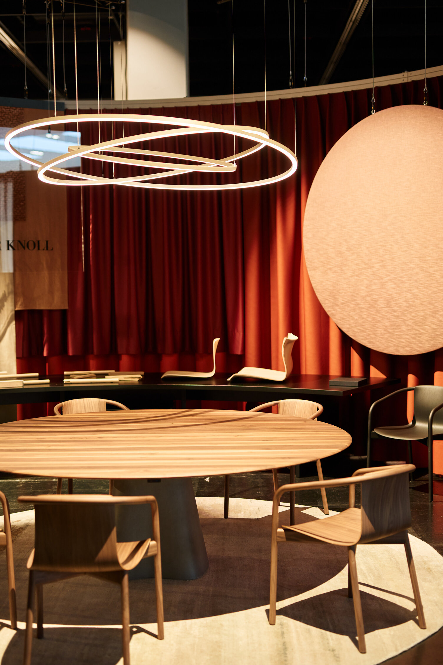 PURE MOTION SLIM über rundem Tisch bei WALTER KNOLL | © Philip Kottlorz Messestand von WALTER KNOLL auf der ORGATEC mit einem runden Holztisch und Holzstühlen darum, darüber schweben drei ineinander verkreuzte Ringleuchten, die PURE MOTION. | © Philip Kottlorz