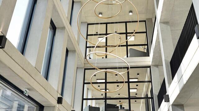 Moderner Eingangsbereich eines architektonisch anspruchvollen Betongebäudes von  Monari. Geschmückt durch schwarze Fensterrahmen. Blick auf die Empfangstheke mit insbesonderem Augenmerk auf den individuellen goldenen Kronleuchter aus 6 SLIM Ringleuchten nach außenstrahlend über der Empfangstheke. | © Monari