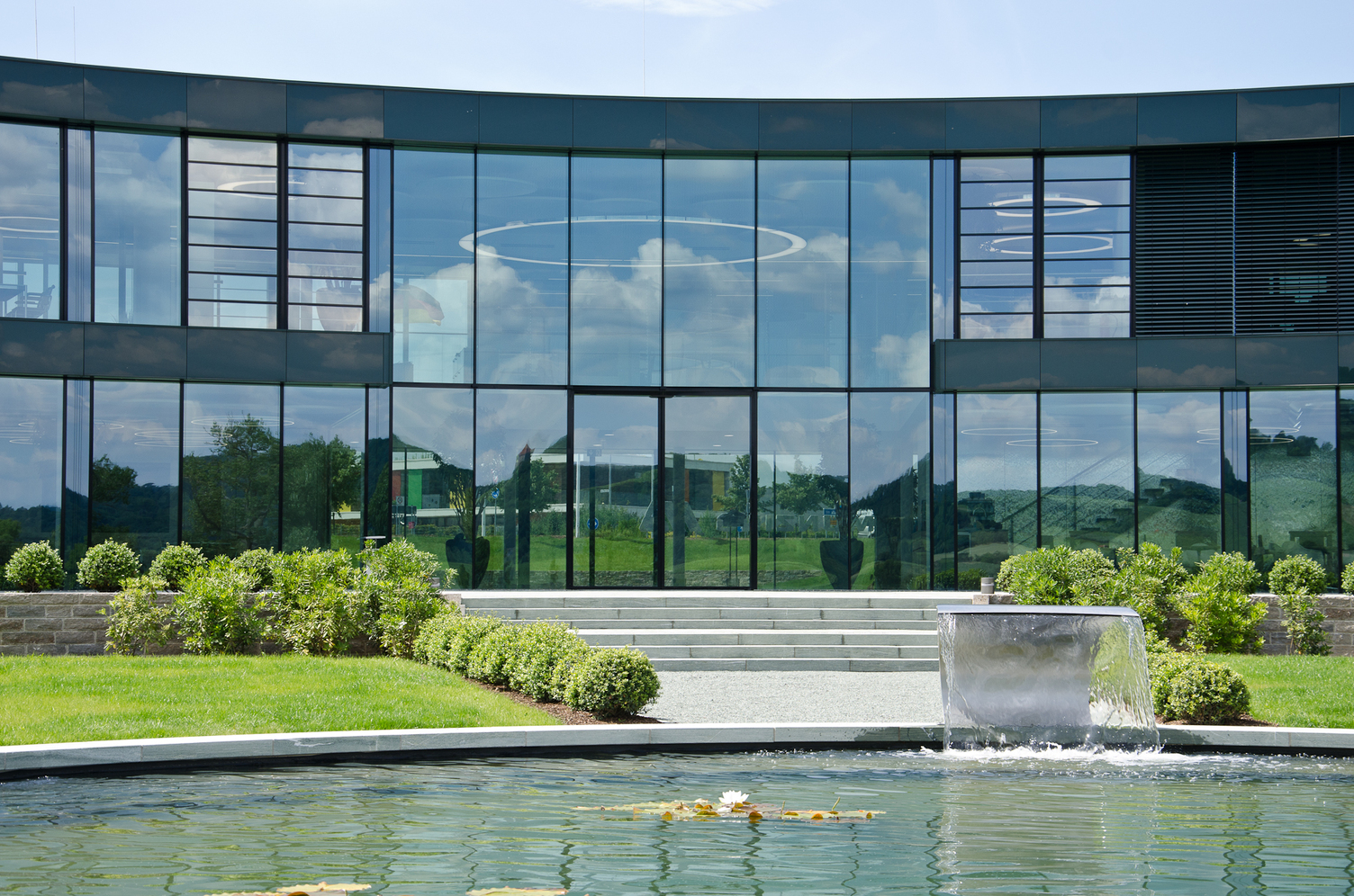 Außenansicht eines modernen Bürokomplexes mit einer Glasfront, wodurch man die mittig angeordnete Anello Leuchte der Firma SATTLER in einem Foyer sehen kann.