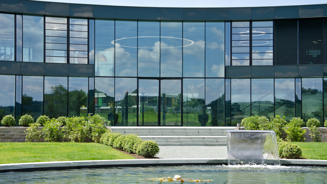 Außenansicht eines modernen Bürokomplexes mit einer Glasfront, wodurch man die mittig angeordnete Anello Leuchte der Firma SATTLER in einem Foyer sehen kann. | © SATTLER | Werbe-und Medienagentur Mohring