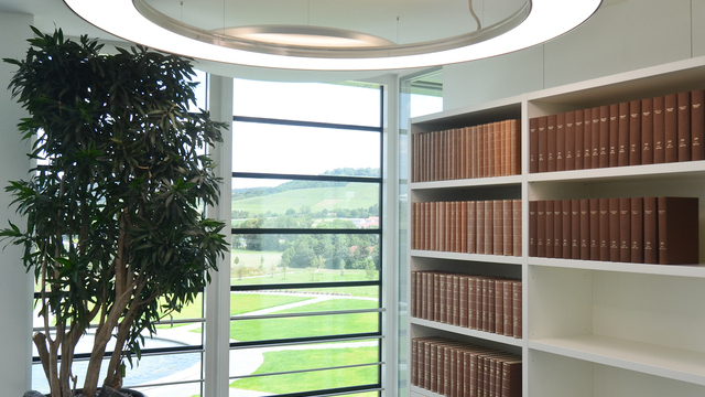 In einer Büroverkehrsfläche mit Bücherregal im Hintergrund wurde esthetisch eine Anello im Durchmesser 2m in Aluminium poliert in Kombination mit Acoustic Panels jeweils von SATTLER eingesetzt. | © SATTLER | Werbe-und Medienagentur Mohring