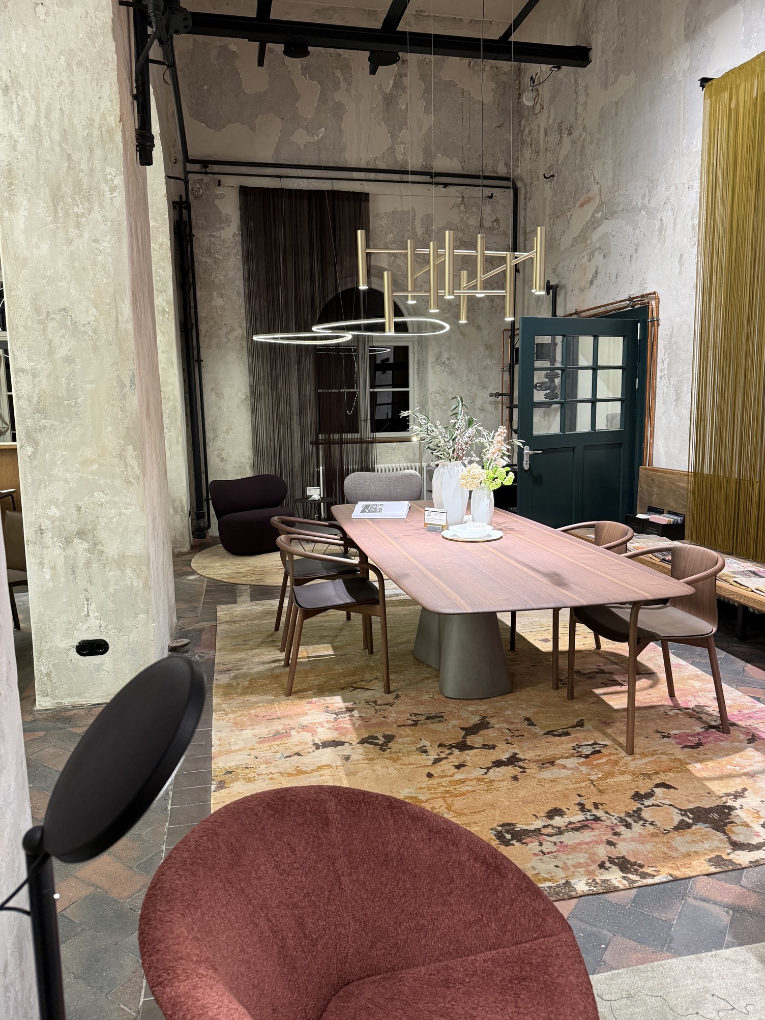 Holz-Esstisch mit Holz-Stühlen von WALTER KNOLL in shabby-chic Raum, über dem Tisch ist ein moderner Kronleuchter EVOLUTION M bestehend aus zylindrischen Elementen in der Oberflächenfarbe Champagner-Gold abgependelt. Im Hintergrund sind abgerundete leuchtende Dreiecksleuchten SLIM DELTA direct zu sehen.