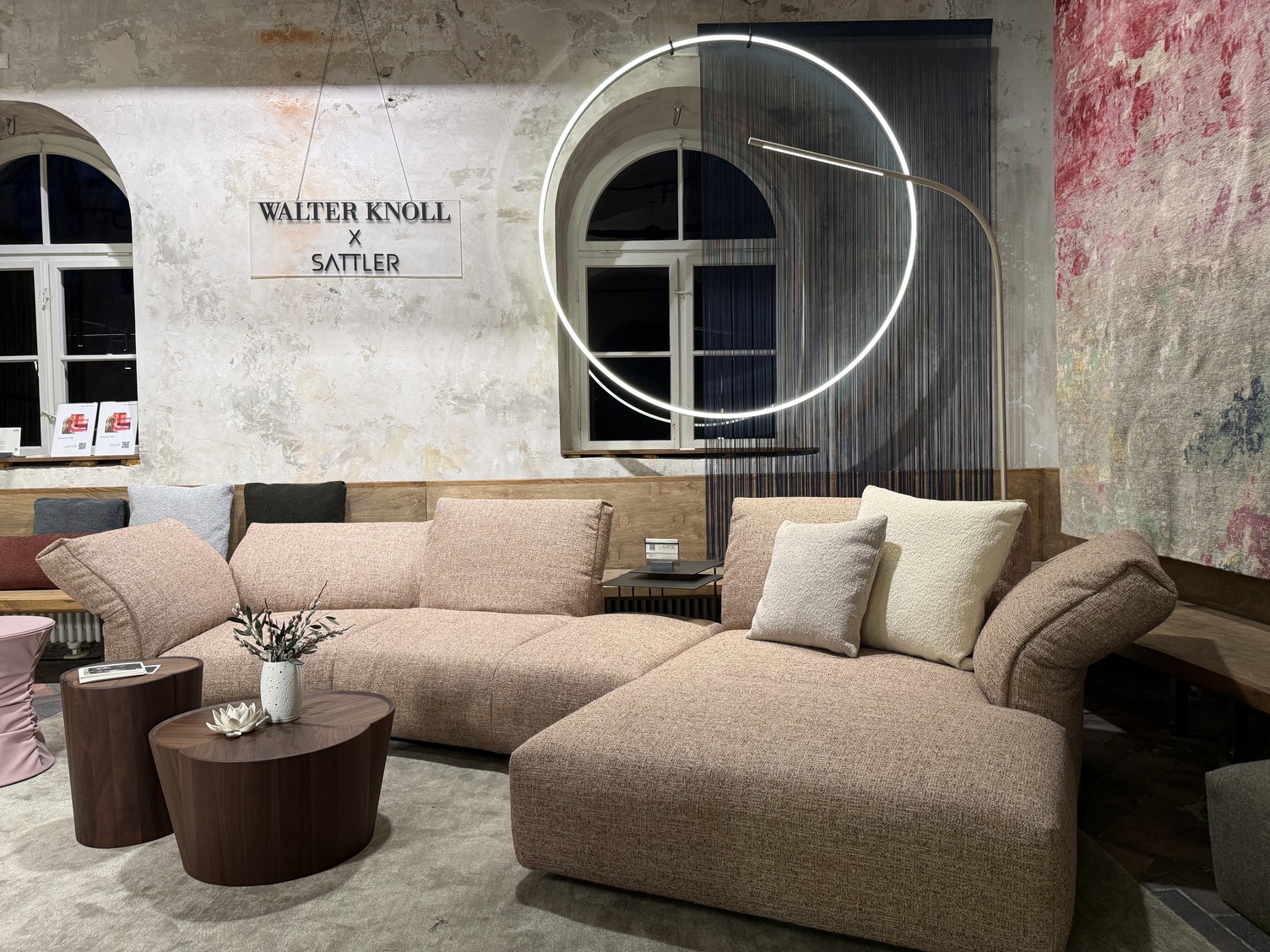 Halbrunde Sofalandschaft von Walter Knoll, im Hintergrund ein Bogenfenster, über dem oberen Teil ist davor eine Runde Ringleuchte vertikal abgependelt, DOPPIO von SATTLER. Hinter dem Sofa steht eine goldene gebogene Stableuchte PALITO FLOOR. An der Wand im Hintergrund stehen die Namen: WALTER KNOLL x SATTLER.