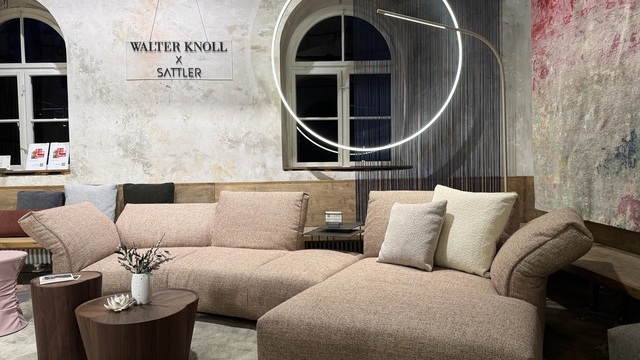 A semi-circular sofa arrangement by Walter Knoll, with an arched window in the background; suspended vertically above the upper section is a circular ring light, DOPPIO by SATTLER. Behind the sofa stands a golden, curved floor lamp, PALITO FLOOR. On the wall in the background are the names: WALTER KNOLL x SATTLER. | © SATTLER