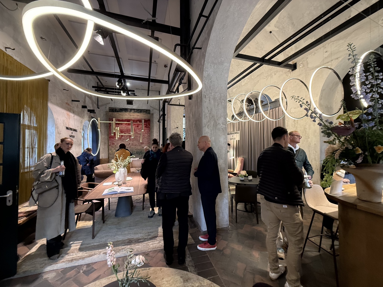 Shabby-chicer Raum mit vielen runden Durchbrüchen, belebt mit mehreren Menschen, die die Leuchten von SATTLER und Möbel von Walter Knoll auf den Munich Design Days 2026 anschauen. Pendelleuchten, Stehleuchten, Kronleuchter und mehr von SATTLER sind zu sehen, arrangiert bei Tischen, Sofas und Sesseln von Walter Knoll.