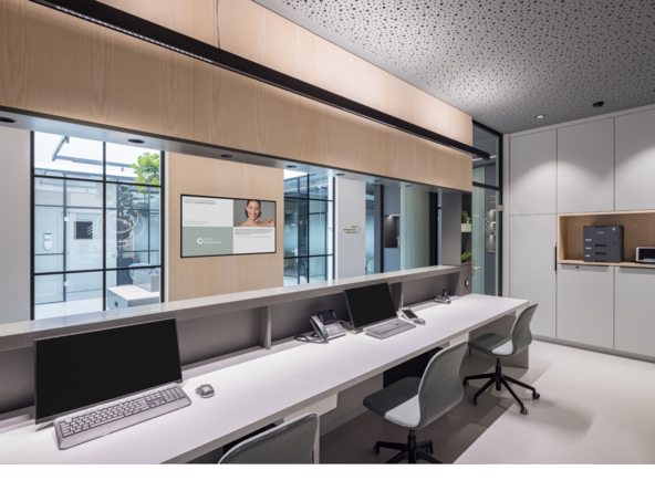 Reception desk of a medical practice with a view from the workstations to the corridor, with glare-free PALADIO LENS linear luminaires above the monitor workstations and BE spotlights on the ceiling by the neighbouring cabinets. | © Lars Gruber