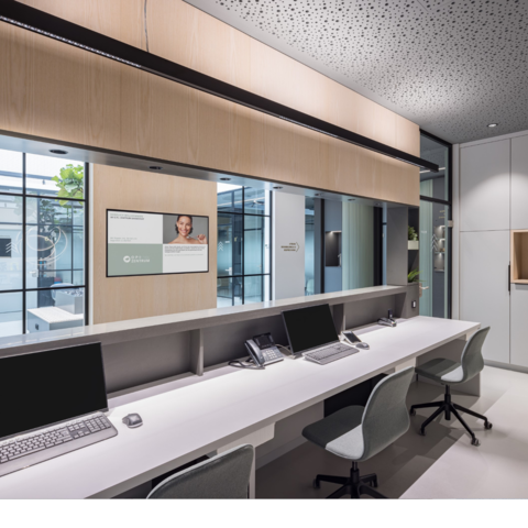 Reception desk of a medical practice with a view from the workstations to the corridor, with glare-free PALADIO LENS linear luminaires above the monitor workstations and BE spotlights on the ceiling by the neighbouring cabinets.