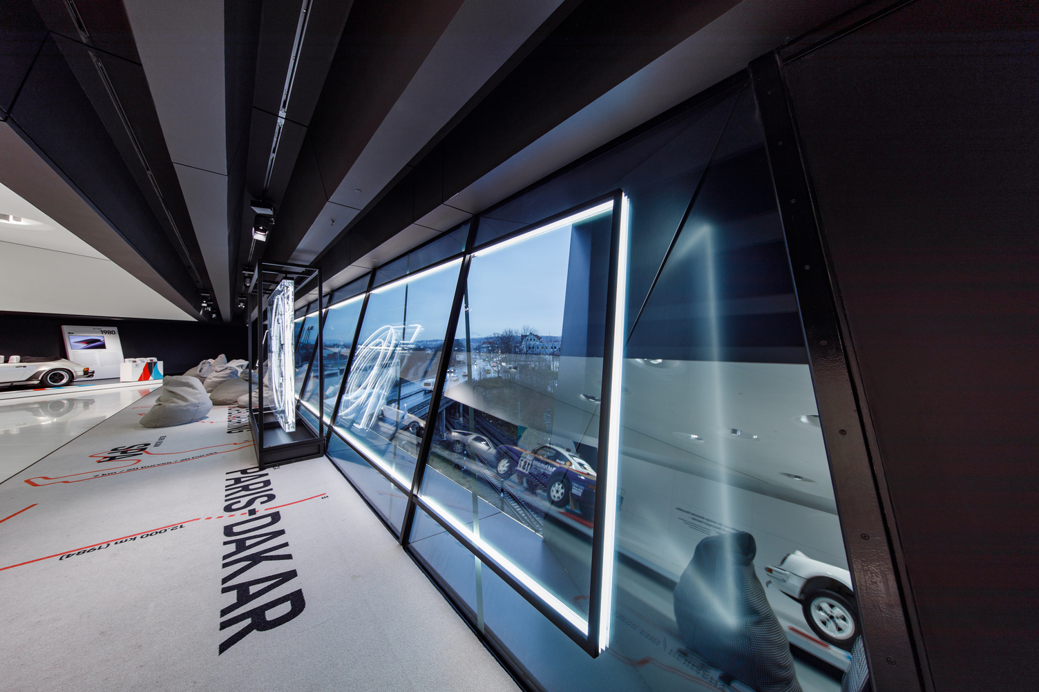 Innenansicht des Porsche Museums in Stuttgart mit der Paris-Dakar Ausstellung und moderner Beleuchtung von Sattler zum 70 jährigen Jubiläum von Porsche Sportwagen. Für die Fenster wurde ein beleuchteter Rahmen als Bbeleuchteter Bilderrahmen mit unsererm CoveLighting in einer Sonderausführung geplant, entwickelt und montiert.