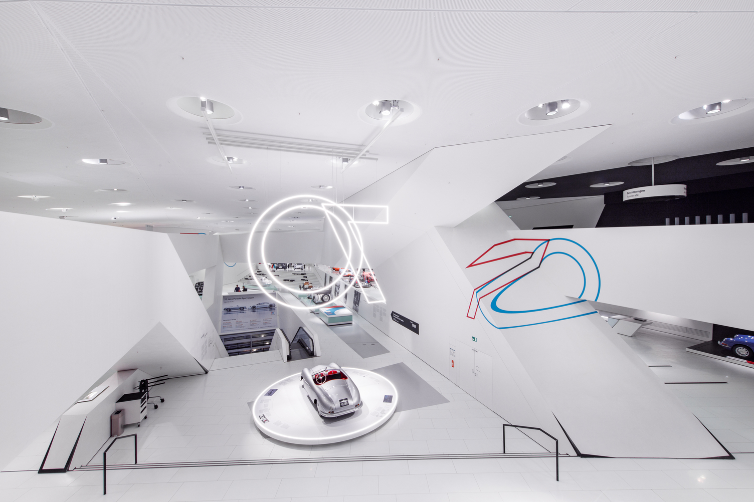 Weite Innenansicht des Porsche Museums zum Jubiläum 70 Jahre Porsche Sportwagen in Stuttgart, die den Ausstellungsraum mit moderner Beleuchtung von Sattler zeigt. Es wurde zu diesem Zweck aus der Leuchtenserie Doppio von SATTLER eine 70 geformt und von der Decke über den Sportwagen ausgestellt. Zum Tragen kamen Doppio Linea und Doppio Rundleuchten, welche jeweils vertikal von der Decke abgependelt wurden um von allen Seiten des Gebäudes im Inneren sichtbar zu sein.