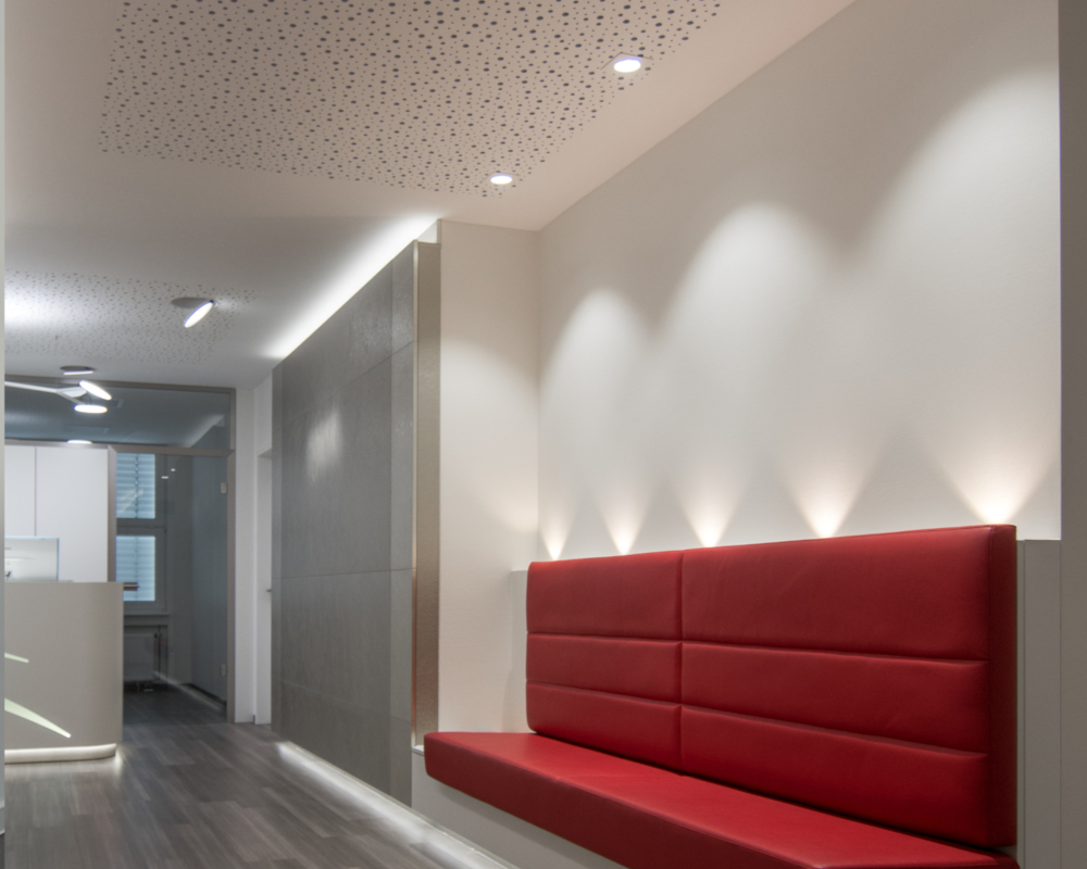 Hallway lighting with BE downlights Practice waiting area in the corridor with a red bench, behind which five beams of light shine upwards, and three spotlights on the ceiling (BE Downlights) shine downwards onto the bench, with the reception desk visible in the background.