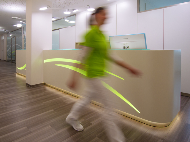 A woman wearing a green T-shirt and white trousers walks past a long white reception desk with green lines integrated into it. AVVENI pendant luminaires with round, flat luminaire heads pointing in different directions hang above the desk.