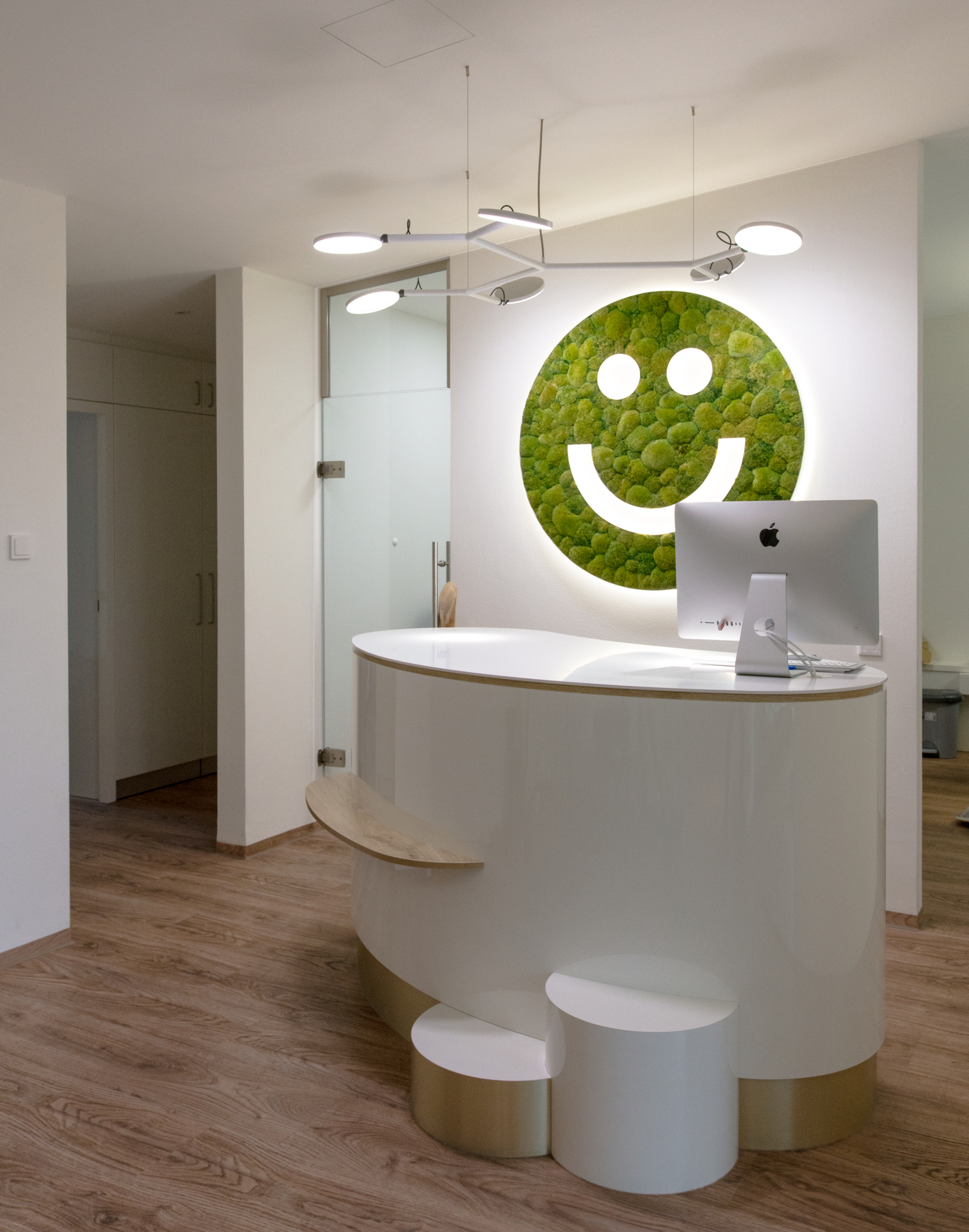 Freundlicher weißer Empfangstresen der Arztpraxis Dr. Veronika Wolff mit einer verzweigten Pendelleuchte mit 6 Leuchtköpfen, AVVENI PENDANT 6, und einem Moos-Smiley an der Wand dahinter, dessen Augen und Mund leuchten (Sonderanfertigung aus LUCE VERDE).
