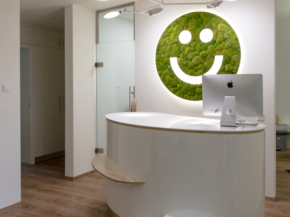 Friendly white reception desk at Dr Veronika Wolff's medical practice with a branched pendant luminaire with 6 light heads, AVVENI PENDANT 6, and a moss smiley on the wall behind it, whose eyes and mouth light up (custom-made from LUCE VERDE).