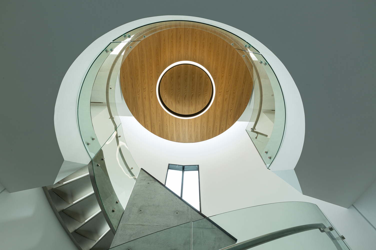 View upwards in a stairwell with a glass handrail and a round outward-radiating luminaire, CIRCOLO INVERSE (diameter 1600), in the centre of the wooden ceiling.