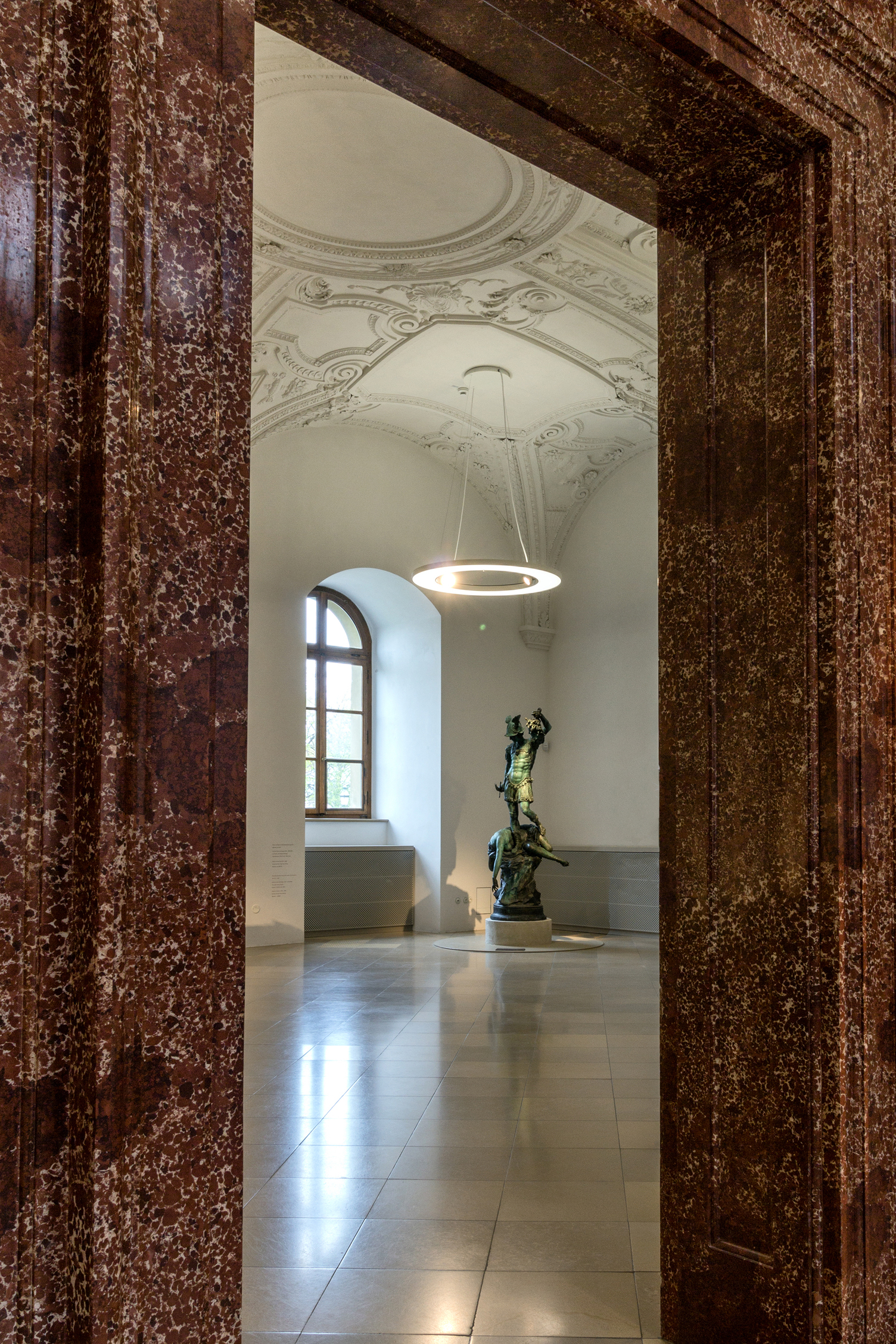Blick durch einen Marmor-Türrahmen in den Bronzesaal der Bayerischen Schlösserverwaltung in der Residenz München, Ausstellungsraum mit Bronze-Skulpturen auf weißen Podesten, von der Stuckdecke mit Gewölbestruktur sind Ringpendelleuchte ANELLO mit integrierten Spots abgependelt, die die Ausstellungsstücke und den Raum ausleuchten. | © Bayerische Schlösserverwaltung, Residenz München