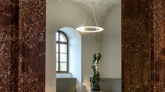 Blick durch einen Marmor-Türrahmen in den Bronzesaal der Bayerischen Schlösserverwaltung in der Residenz München, Ausstellungsraum mit Bronze-Skulpturen auf weißen Podesten, von der Stuckdecke mit Gewölbestruktur sind Ringpendelleuchte ANELLO mit integrierten Spots abgependelt, die die Ausstellungsstücke und den Raum ausleuchten. | © Bayerische Schlösserverwaltung, Residenz München