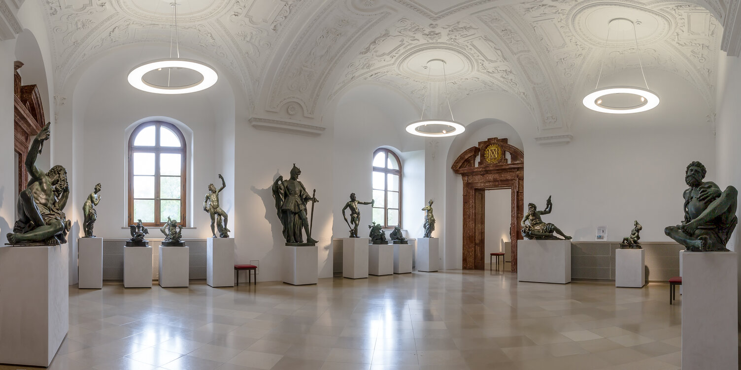 ANELLO Ringleuchten mit Spots in den Bronzesälen der Residenz München | © Bayerische Schlösserverwaltung, Residenz München Bronzesaal der Bayerischen Schlösserverwaltung in der Residenz München, Ausstellungsraum mit Bronze-Skulpturen auf weißen Podesten, von der Stuckdecke mit Gewölbestruktur sind Ringpendelleuchte ANELLO mit integrierten Spots abgependelt, die die Ausstellungsstücke und den Raum ausleuchten. | © Bayerische Schlösserverwaltung, Residenz München