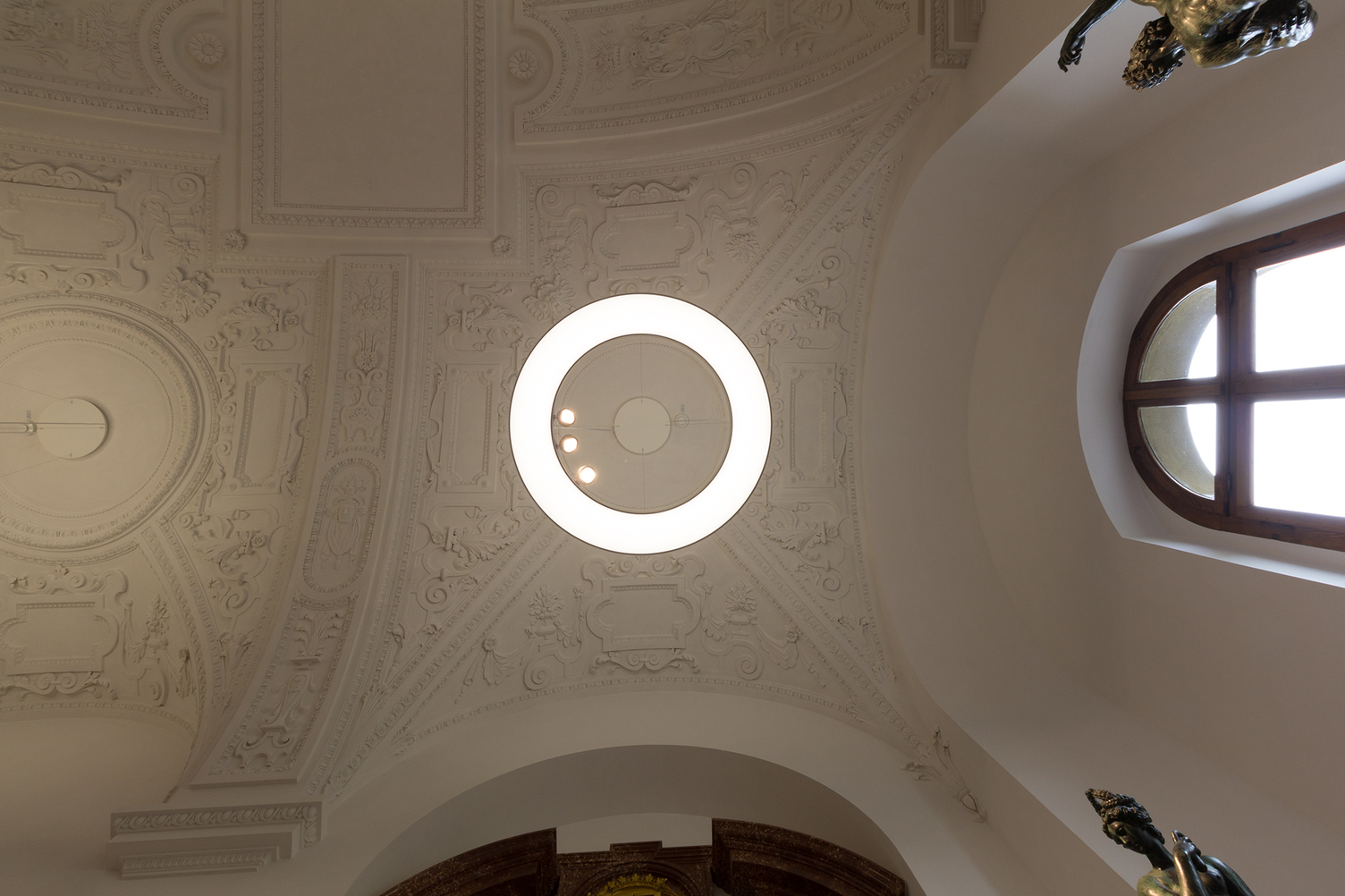 Blick von unten auf eine großformatige Pendel-Ringleuchte ANELLO, die gleichzeitig die Gewölbe-Stuck-Decke & die Exponate beleuchtet. | © Bayerische Schlösserverwaltung, Residenz München