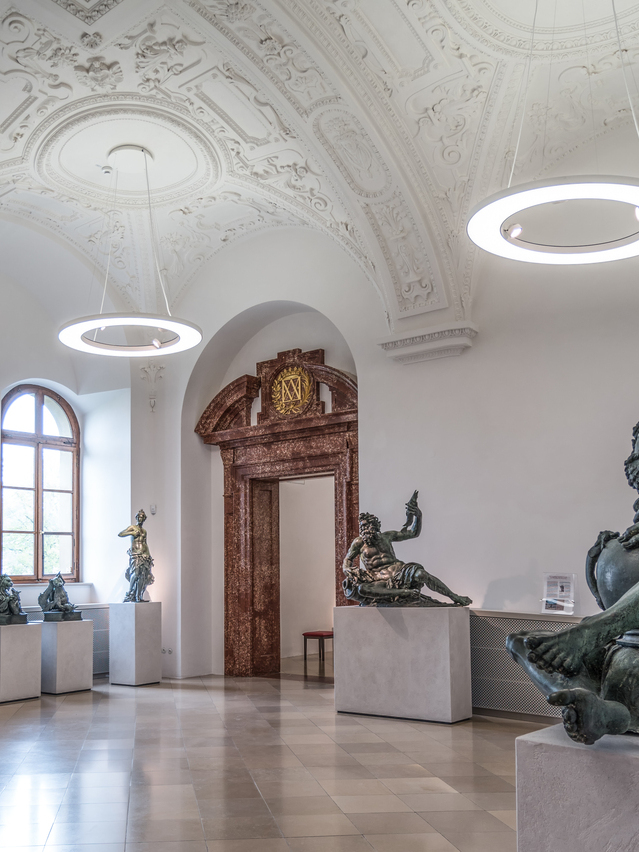 Bronzesaal der Bayerischen Schlösserverwaltung in der Residenz München, Ausstellungsraum mit Bronze-Skulpturen auf weißen Podesten, von der Stuckdecke mit Gewölbestruktur sind Ringpendelleuchte ANELLO mit integrierten Spots abgependelt, die die Ausstellungsstücke und den Raum ausleuchten. | © Bayerische Schlösserverwaltung, Residenz München
