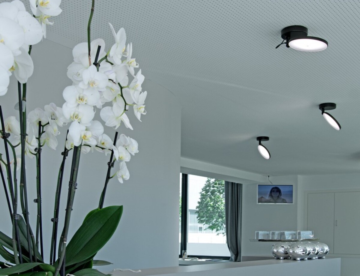 Weiße geteilte lineare Empfangstheke im Friseursalon Mentes. Links steht eine weiße Orchidee auf dem Tresen, an der weißen Lochdecke darüber sind entlang der Thekenlänge einzelne schwarze AVVENI SINGLE, kleine Anbauleuchten mit beweglichen runden flachen Leuchtenköpfen, die in verschiedene Richtungen ausgerichtet sind.