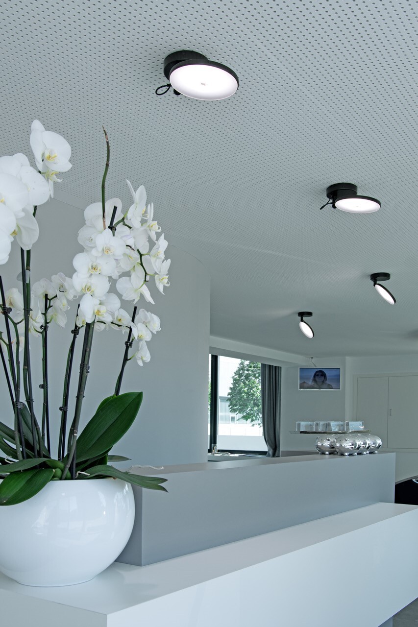 Weiße geteilte lineare Empfangstheke im Friseursalon Mentes. Links steht eine weiße Orchidee auf dem Tresen, an der weißen Lochdecke darüber sind entlang der Thekenlänge einzelne schwarze AVVENI SINGLE, kleine Anbauleuchten mit beweglichen runden flachen Leuchtenköpfen, die in verschiedene Richtungen ausgerichtet sind.