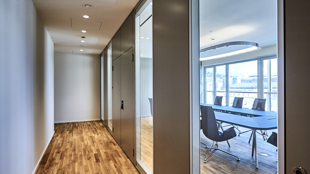 Flur in einem moderenen Büro mit Stäbchen Parkett, der Flur wird von kleinen runden Einbauspots, BE2-Leuchten, erhellt. Man hat Einblick durch eine Glasfront in einen angrenzenden Besprechungsraum mit Tisch, Stühlen und rechteckigen Ringleuchte SOFT SQUARE. | © Fotografie SUL – Sven Lorenz